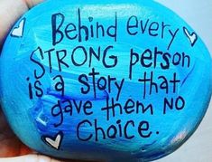 a hand holding a blue painted rock with writing on it that says, behind every strong person is a story that gave them no choice