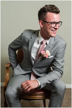 a man in a gray suit and pink tie sitting on a chair smiling at the camera