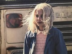 a woman with blonde hair standing in front of an old bus and looking at the camera