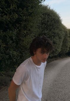 a young man standing on the side of a road next to trees and bushes in front of him