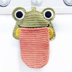 a crocheted frog oven mitt hanging on the wall with its tongue out