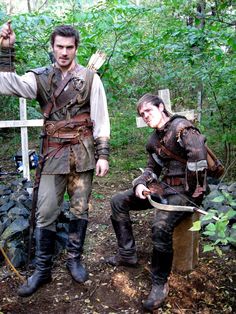 two men dressed in medieval clothing standing next to each other on a forest path with trees and bushes behind them