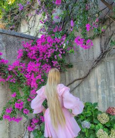 a blonde haired woman with long hair standing in front of purple flowers and greenery