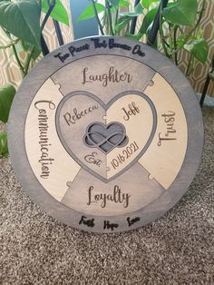 a wooden plaque with two hearts and names on it, sitting in front of a potted plant