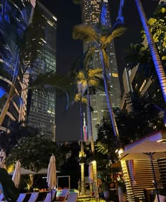 palm trees and lounge chairs are lit up in the city at night with skyscrapers behind them