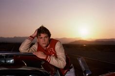 a young man leaning on the hood of a red sports car in front of a sunset