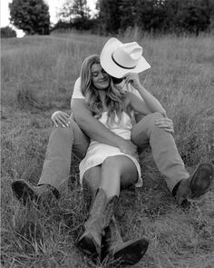 black and white photograph of couple sitting in field with cowboy hat on their head, hugging each other