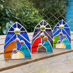 three colorful stained glass pieces sitting on top of a wooden table in front of some bushes