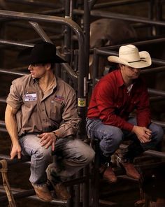 two men in cowboy hats are sitting on some metal bars and one is wearing a red shirt