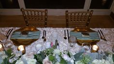 the table is set with silverware, plates and flowers in front of two gold chairs