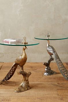 two metal peacock figurines sitting on top of a wooden floor next to a glass table