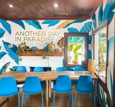 a dining room with blue chairs and a large painting on the wall that says another day in paradise