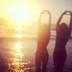 two people standing in the water making a heart shape with their hands while the sun is setting