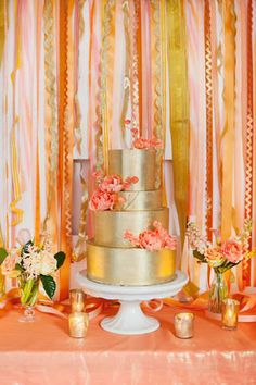 a wedding cake with pink and gold flowers on it sitting on a table in front of an orange curtain