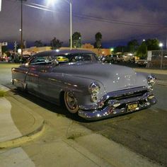 an old car is parked on the side of the road at night with other cars in the background