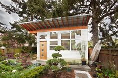 a small white house surrounded by trees and plants in front of a fenced yard