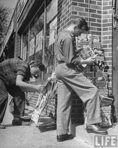 two men are looking at newspapers on the sidewalk in front of a brick storefront