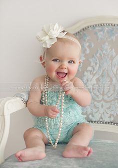 a baby with pearls and a flower in her hair sitting on a chair smiling at the camera