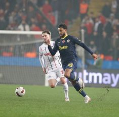 two men playing soccer on a field with people watching from the stands in the background