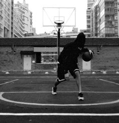 black and white photograph of a man playing basketball on an outdoor court with buildings in the background