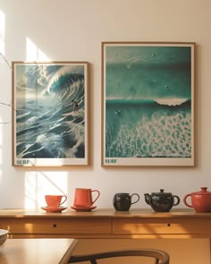 two framed pictures hang on the wall above a table with cups and teapots