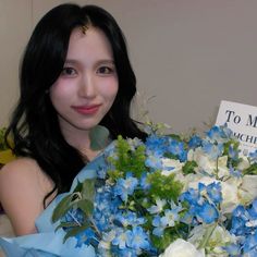 a woman holding a bouquet of blue and white flowers in her hands with a sign on the wall behind her