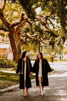 two women in graduation gowns walking down the street holding hands and looking into each other's eyes