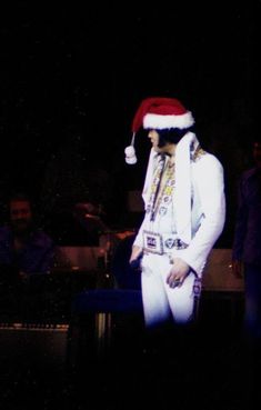 a man wearing a santa hat and white suit on stage with other people in the background