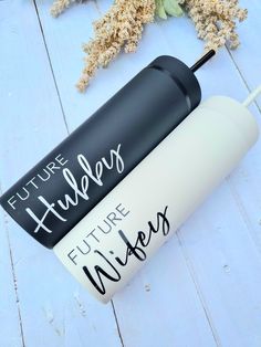 two white and black travel mugs sitting on top of a wooden table next to dried flowers