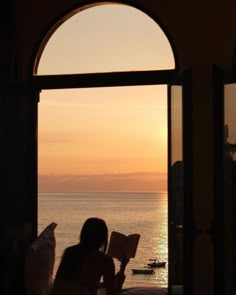 a woman sitting on a bed reading a book in front of an open window at sunset