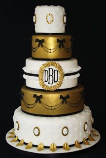 a three tiered wedding cake decorated with gold and black ribbon, bows and monograms