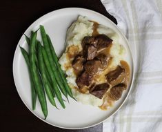 a white plate topped with meat and mashed potatoes covered in gravy next to green beans