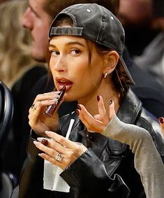 a woman in black jacket and hat eating food at a basketball game with other people watching