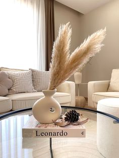 a glass table topped with a white vase filled with dry grass and a pine cone