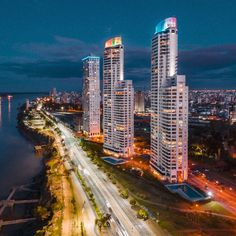 an aerial view of some very tall buildings by the water at night with lights on