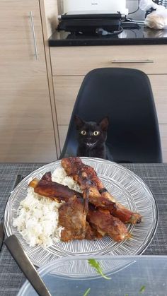 a black cat sitting behind a plate of food on a table with utensils