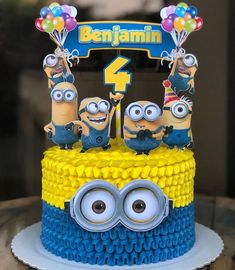 a blue and yellow birthday cake with three minion characters on the bottom tier, surrounded by balloons