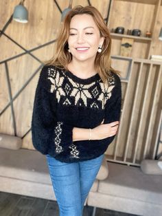 a woman standing in front of a wooden wall wearing a black sweater and blue jeans