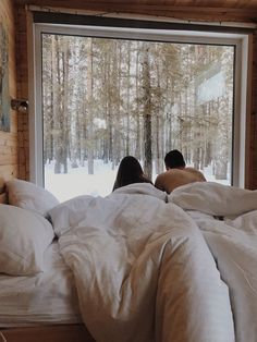 two people are sitting in bed looking out the window at the snow covered trees outside