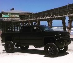 a black truck parked on top of a sandy beach next to the ocean with a pier in the background