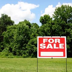 a red for sale sign sitting in the middle of a grass field next to trees