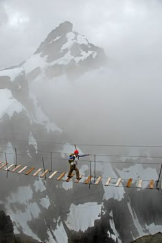 a man walking across a bridge in front of a snow covered mountain with skis on it