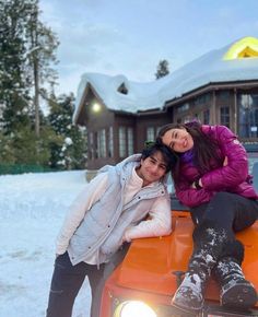 two women sitting on the hood of an orange truck in front of a house covered in snow