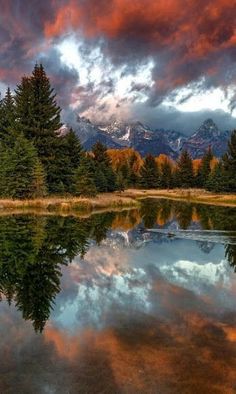 a lake surrounded by trees and mountains under a cloudy sky with red, orange, and blue clouds