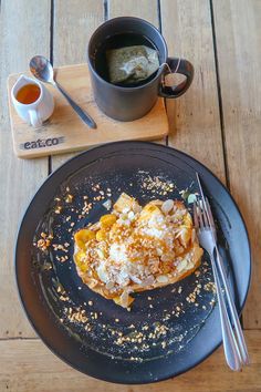 a black plate topped with food on top of a wooden table next to a cup of coffee