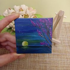 a person holding up a small piece of art that looks like a tree with purple flowers on it