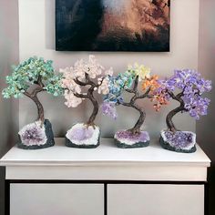 three bonsai trees are displayed on a white table with purple, green and blue rocks