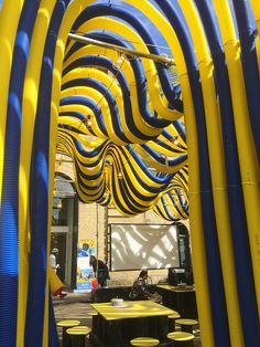 an outdoor area with yellow and blue tubes on the ceiling, tables and stools