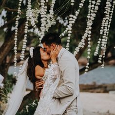 a bride and groom kissing under a tree with white flowers hanging from it's branches