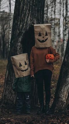 two children standing in the woods with bags on their heads, one holding an orange pumpkin